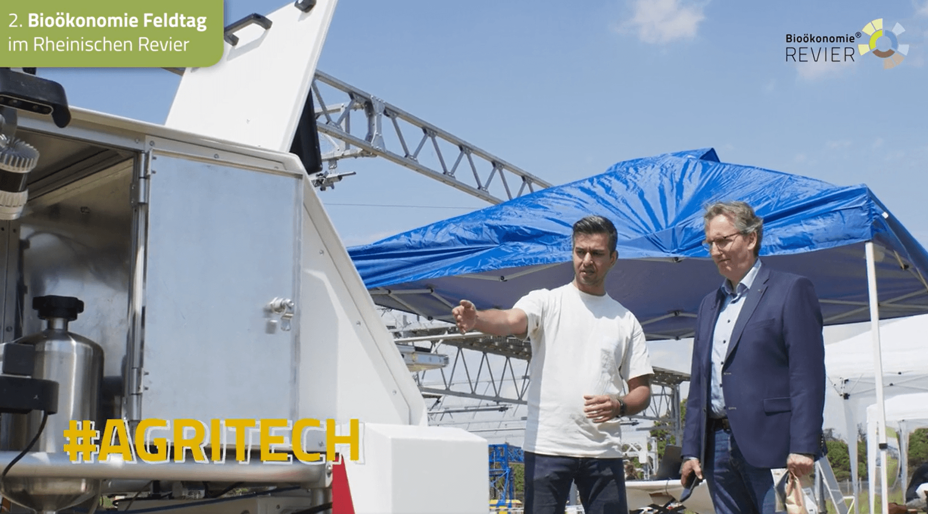Two men talking beside the DynamoBot robot under a blue tent at the Bioökonomie field day in the Rheinisches Revier.