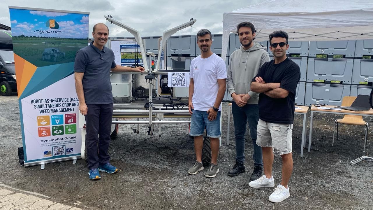 Four DynamoBot team members in front of the robot and a banner about robot-as-a-service weed management with UN SDG icons.