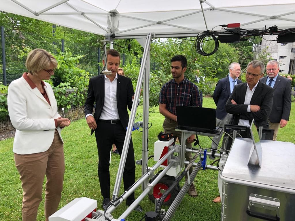 DynamoBot robot with a mounted laptop demonstrated under a tent to a group of officials in business attire.