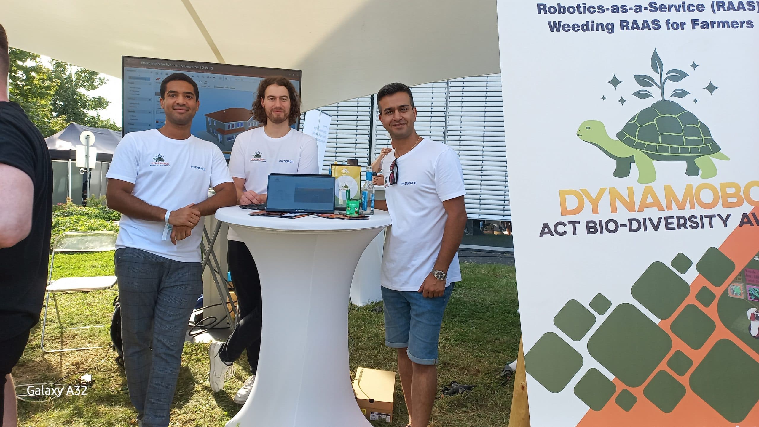 Three DynamoBot team members at a standing table by a booth banner reading Robotics-as-a-Service weeding for farmers.