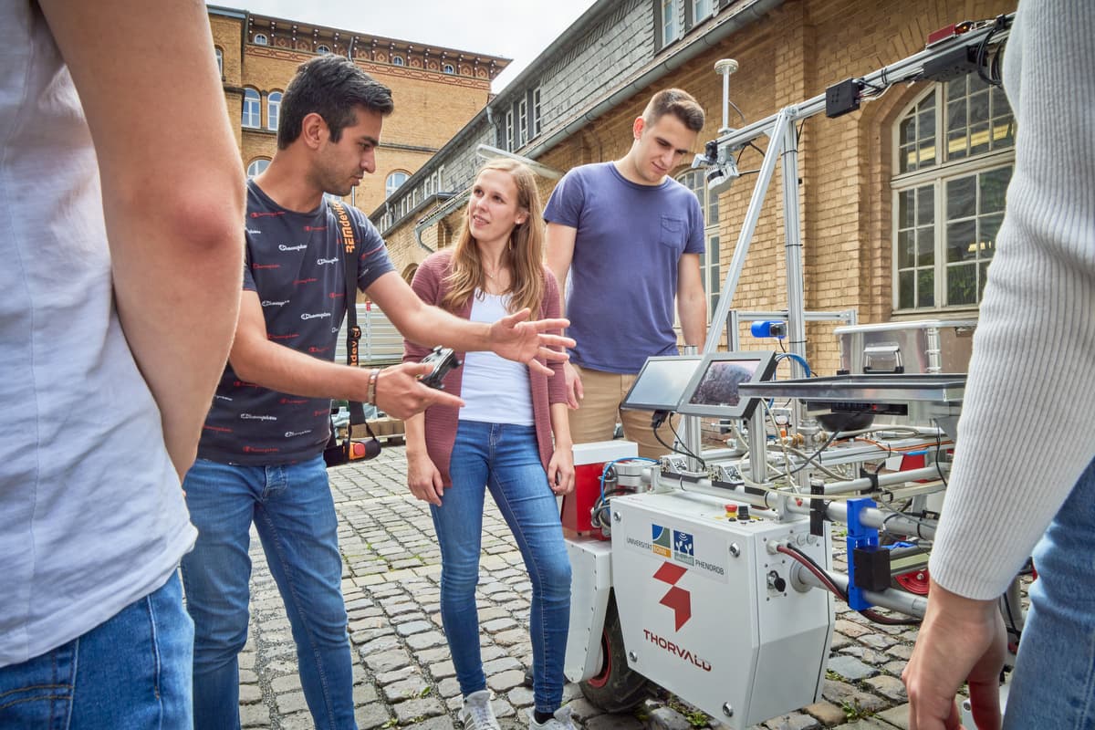 Students around the DynamoBot robot in a cobblestone courtyard while a team member explains it.