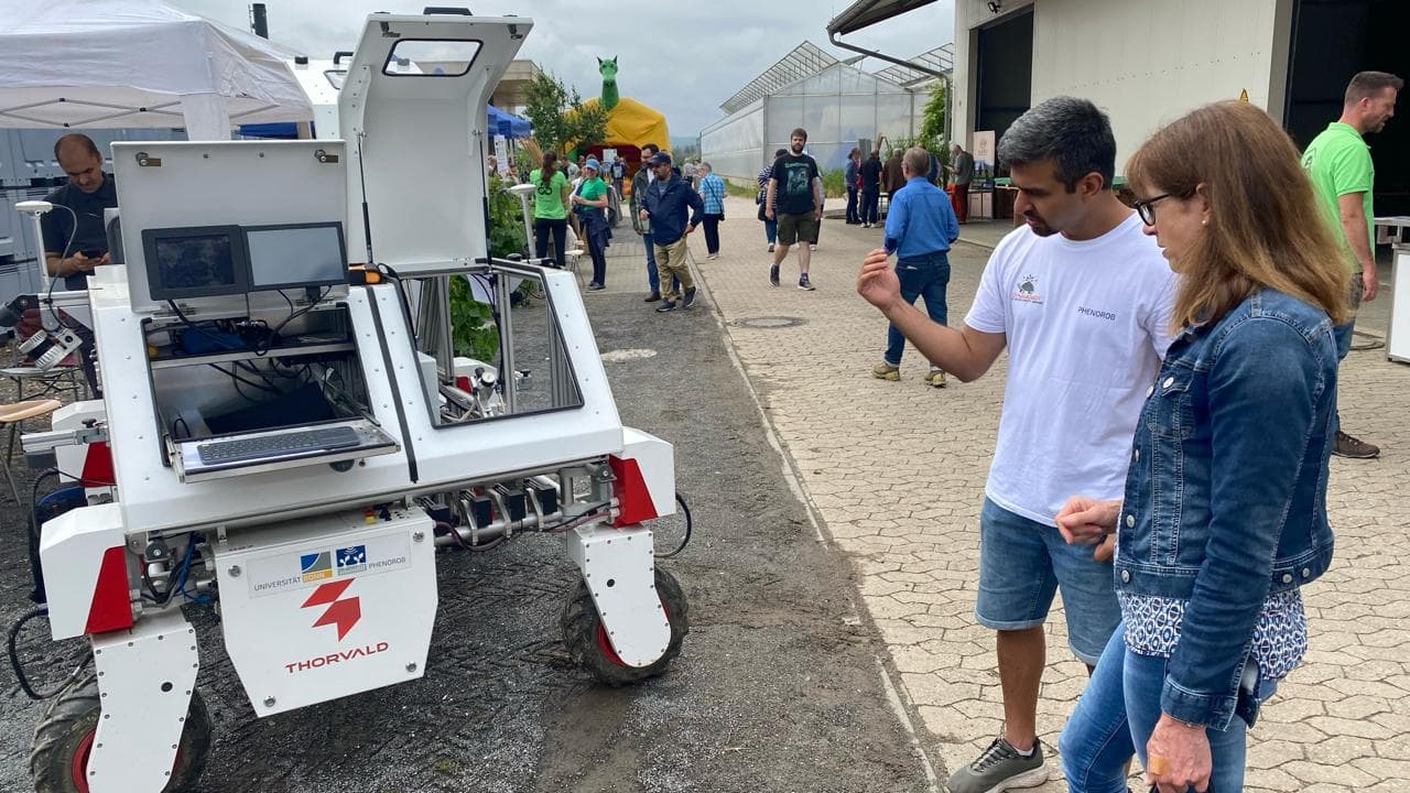 DynamoBot weeding robot on a paved path at an agricultural field day as two visitors look at it.