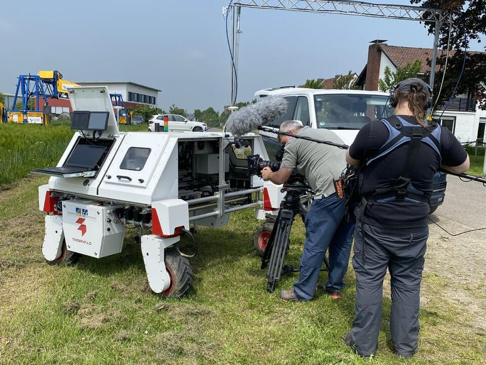 White DynamoBot weeding robot on grass while a film crew with camera and boom mic records it outdoors.